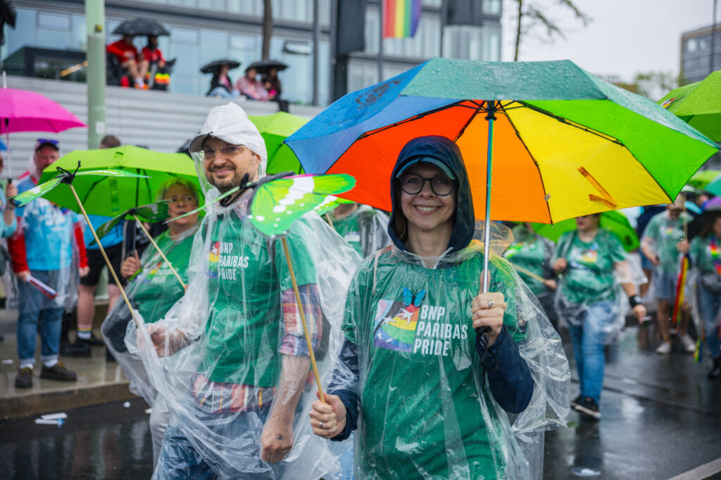 Teilnehmer einer Pride-Parade tragen grüne BNP Paribas Pride T-Shirts und durchsichtige Regenponchos, halten bunte Regenschirme bei Regen.