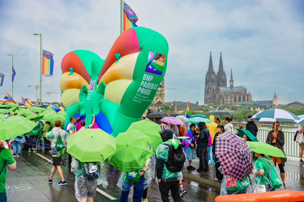 Menschen mit grünen Regenschirmen und bunten BNP Paribas Pride Luftballons auf einer Brücke vor dem Kölner Dom bei Regen