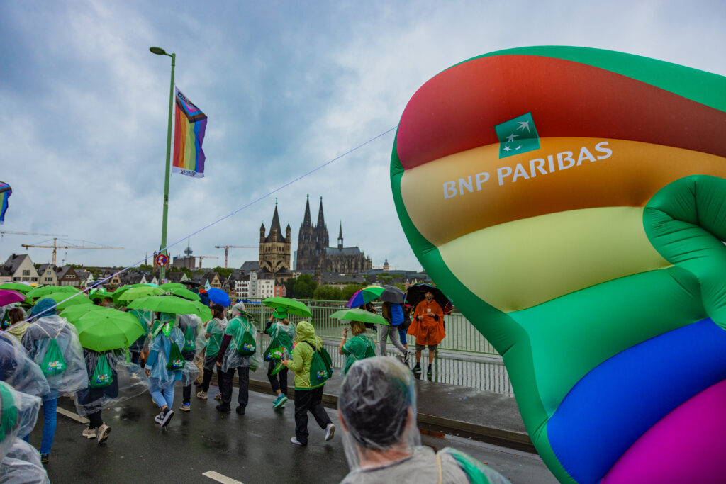 Menschen mit grünen Regenschirmen und Regenponchos auf einer Brücke, im Vordergrund ein großer bunter BNP Paribas Luftballon, im Hintergrund der Kölner Dom und Regenbogenfahnen